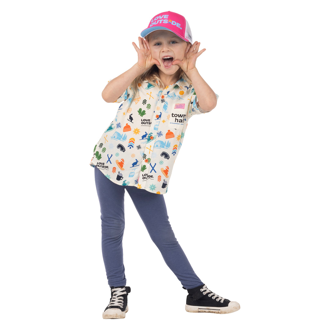 Child wearing Town Hall Kids Cabana Shirt in Vanilla with colorful ski-town print and a pink Love Outside cap.